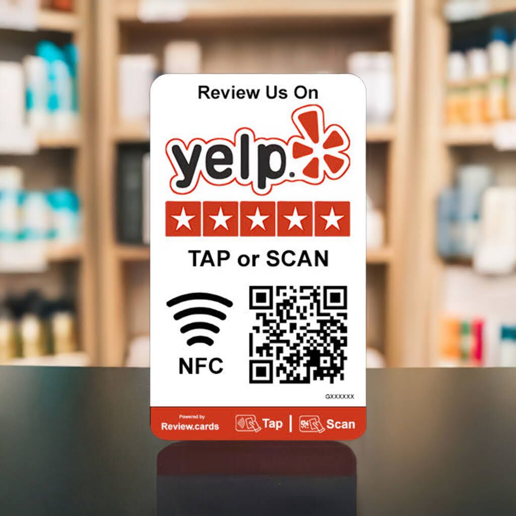A card displayed on a counter has a Yelp logo, five-star icons, and a QR code. The text reads, "Review Us On Yelp," followed by "TAP or SCAN" and an NFC symbol. The background shows blurred shelves with various products. Remember to use Yelp Tap or Scan Smart Review Cards - Free Delivery for your review!