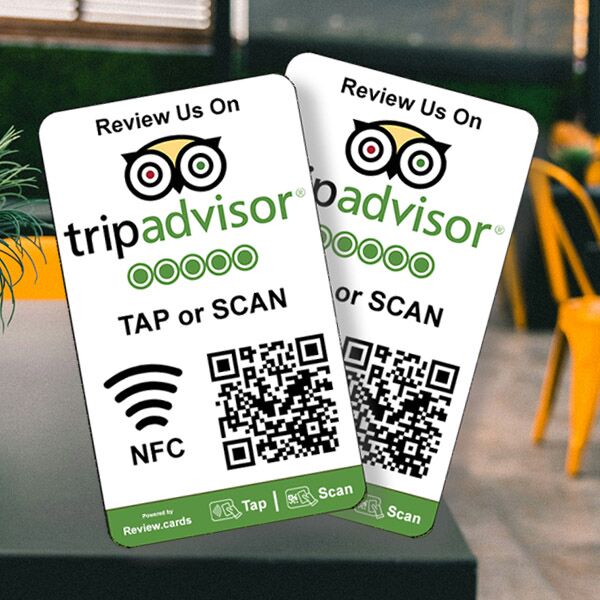 Two "Hospitality Bundle 1" table tents are placed on a table. Each tent features the TripAdvisor logo, a 5-star rating graphic, and instructions to use NFC or a QR code to "Review Us On TripAdvisor." A brightly lit café serves as the backdrop.