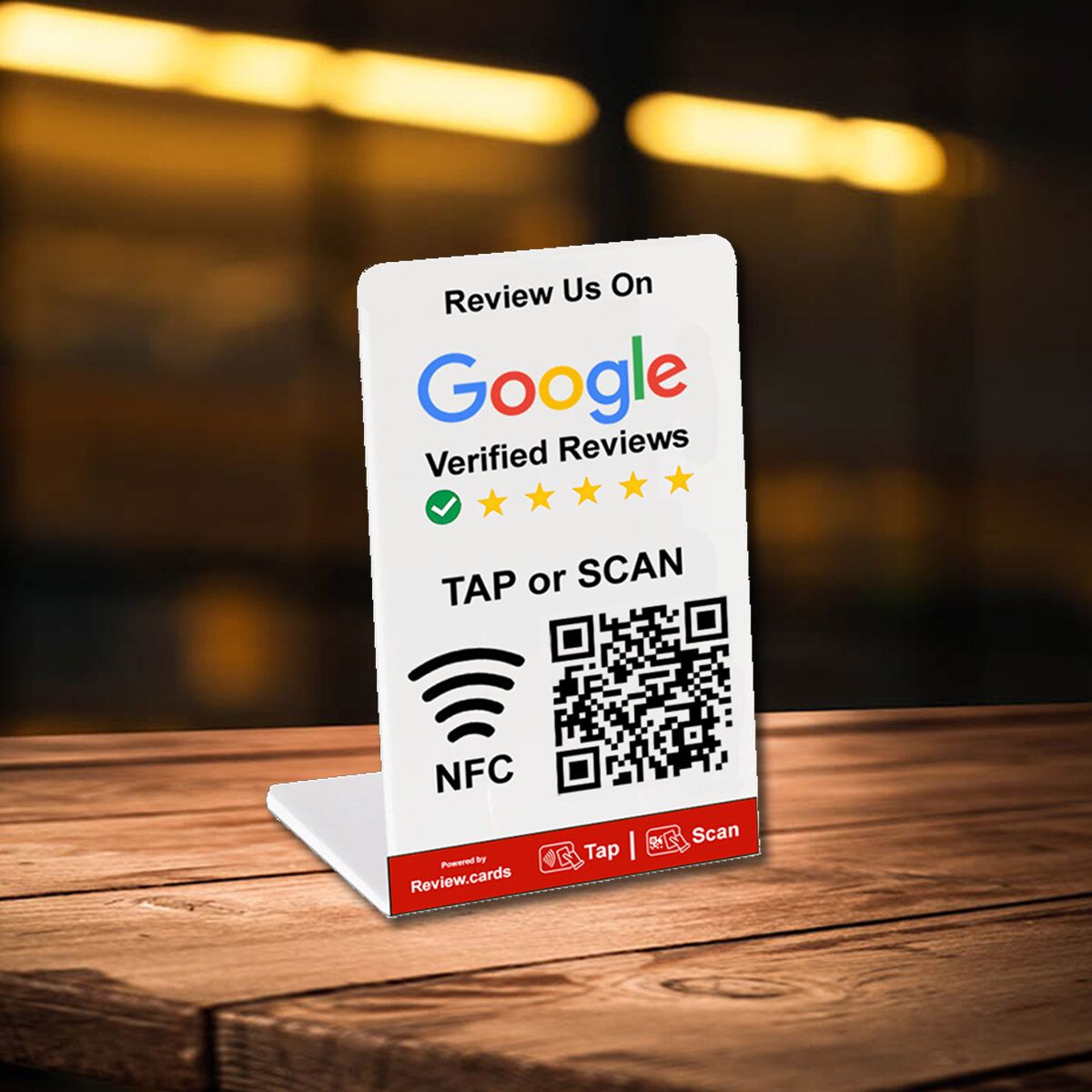 A Hospitality Bundle 1-themed table tent card promoting Google Verified Reviews sits on a wooden table. Featuring a QR code, NFC symbol, and instructions to "TAP or SCAN," it is brightly lit with a blurred background. The bottom reads "Powered by Review.cards" with Tap and Scan icons.