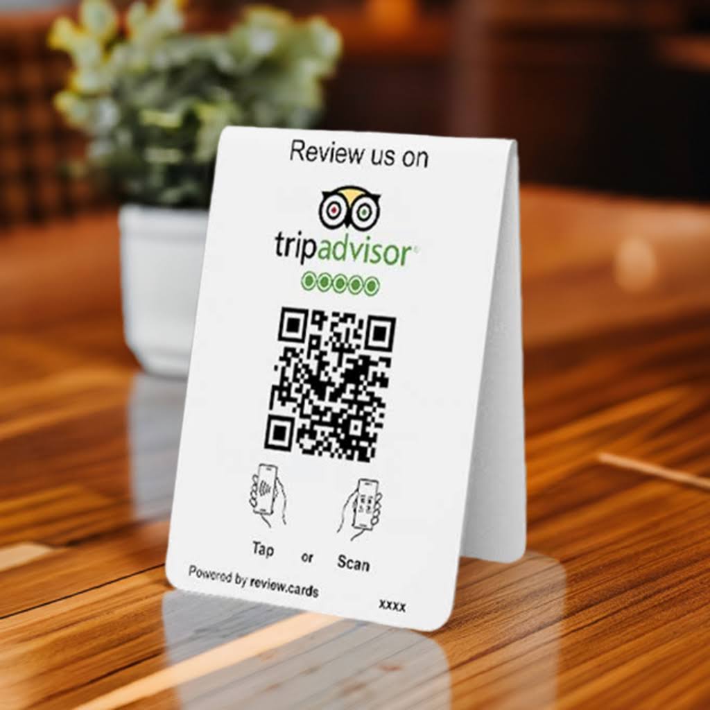 A small table tent card from the Hospitality Bundle 1 sits on a wooden surface, prompting customers to "Review us on TripAdvisor." It features a QR code in the center with illustrations indicating to "Tap or Scan." A potted plant is blurred in the background. This innovative card is powered by review.cards.