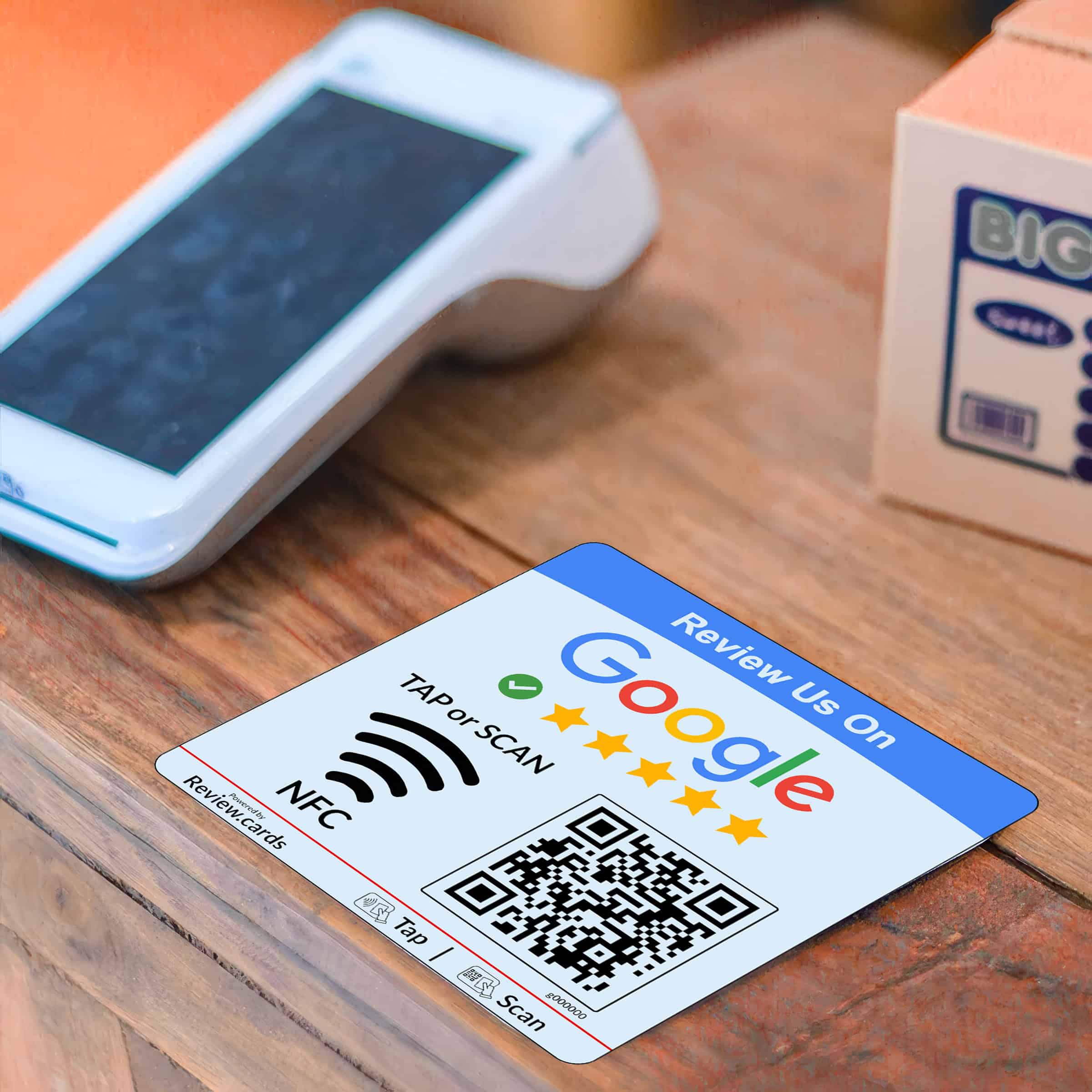A card sits on a wooden counter next to a card reader and a cardboard box. The card, similar in convenience to Google NFC Review Plates, displays a QR code and instructions to "Review Us On Google" with a graphical representation of a five-star review. It also features NFC and tap-to-scan symbols for easy access, ensuring user-friendliness.