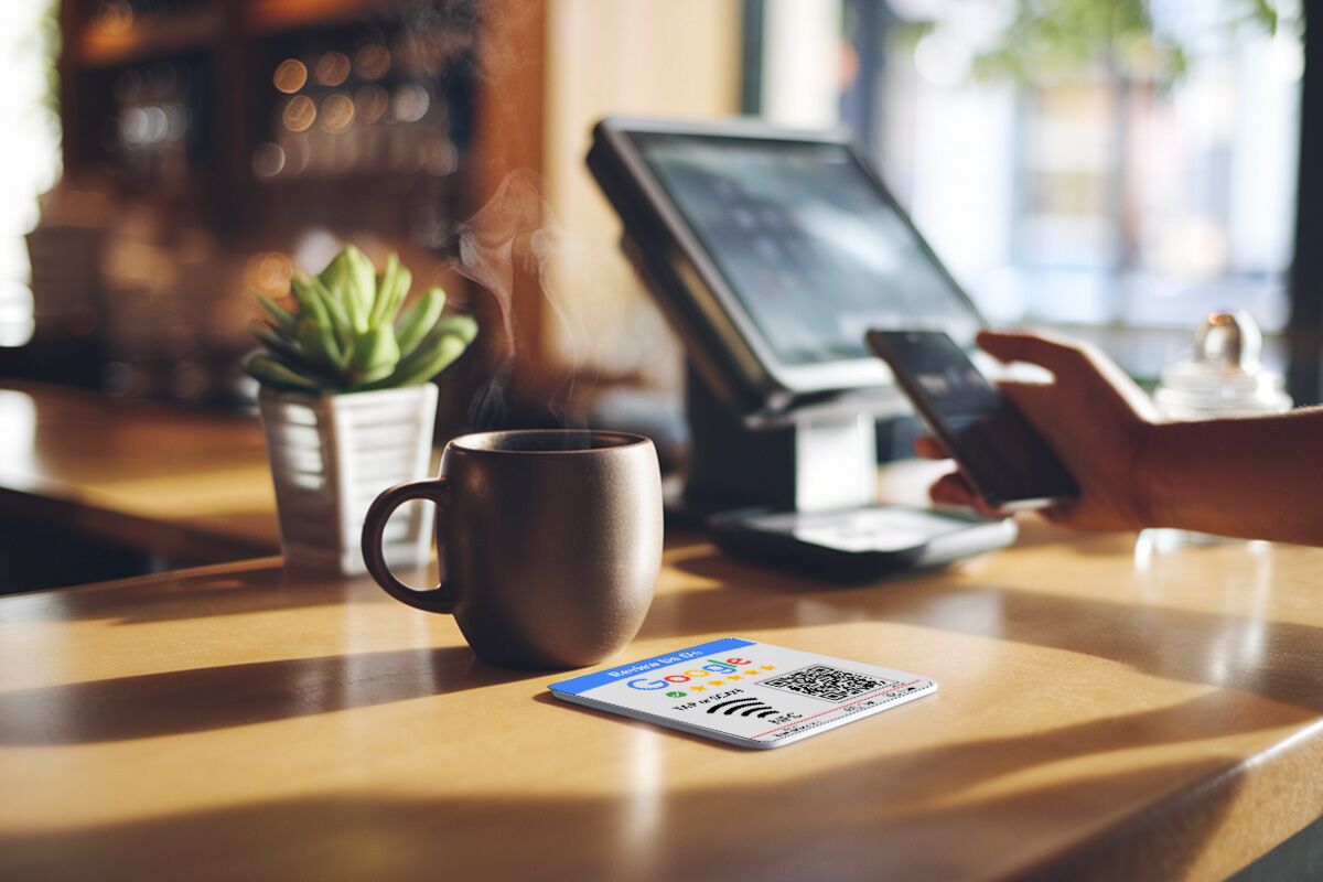 A person holding a smartphone hovers it over a Google Pay QR code card on a café counter. Nearby, a steaming coffee mug and small potted plant accent the scene, while in the blurred background, part of the café interior and Google NFC Review Plates payment terminal can be glimpsed.