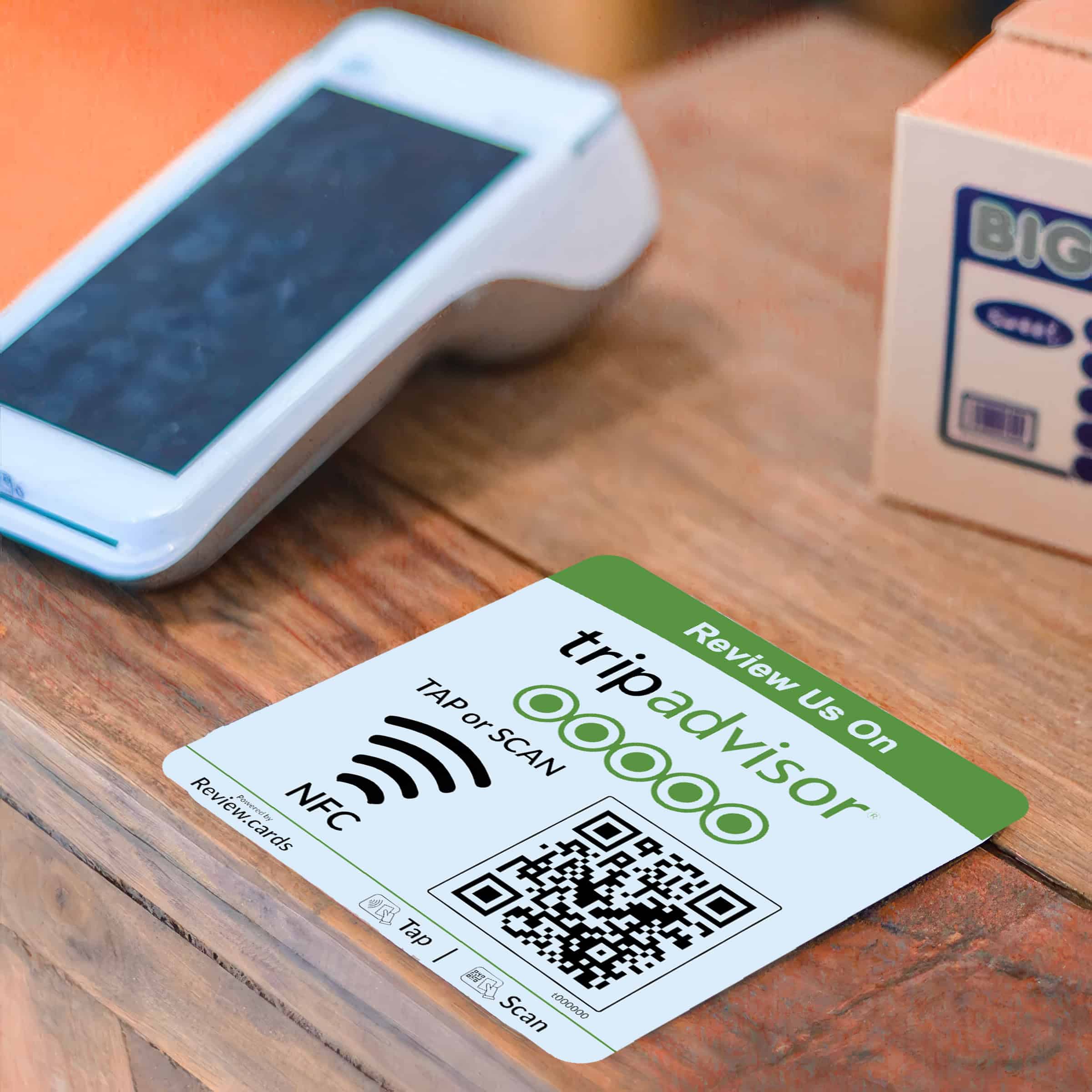 A TripAdvisor NFC Review Plate on a wooden surface displays a QR code, an NFC symbol, and the instruction "Tap or Scan," prompting customers to leave a review. Nearby, there is a smartphone and a point-of-sale terminal. These plates are perfect for gathering quick customer feedback.